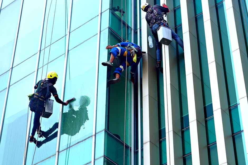 Three window cleaners wearing safety helmets and harnesses are performing surface cleaning on the glass facade of a modern commercial building in Shoreditch. They are using squeegees and cloths to wip