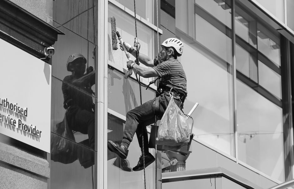 Three window cleaners wearing safety helmets and harnesses are performing surface cleaning on the glass facade of a modern commercial building in Shoreditch. They are using squeegees and cloths to wip