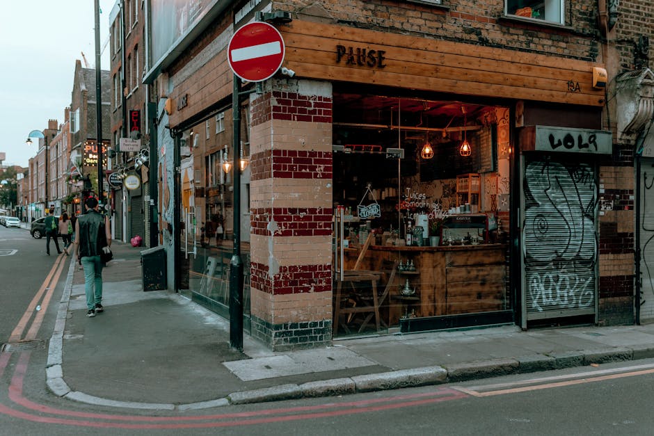 Inside a bus heading through Shoreditch, London, showing the view through the front windshield of a street with brick and modern buildings, trees lining the sidewalk, and a few pedestrians crossing. T